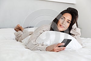 Photo of mature woman 30s using mobile phone, while lying in bed with white linen in bright room