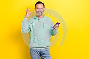Photo of middle age optimistic man brunet hair wear stylish khaki sweater show okey sign convenient phone isolated on