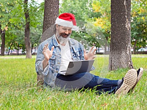 Photo of a middle aged man with a laptop in the park. He sitting on the grass in Santa Hat and communicates with