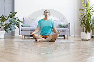 Photo of a middle aged man practicing yoga at home. He sits in the lotus position with headphones and listens to
