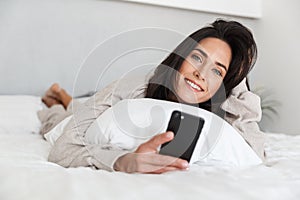 Photo of middle-aged woman 30s using mobile phone, while lying in bed with white linen in bright room