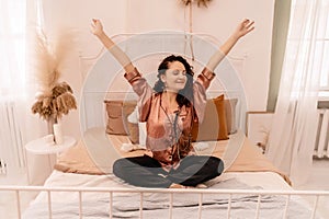 Photo of a middle aged woman in pajamas stretching her arms and smiling while sitting on the bed after sleeping or