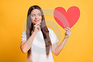 Photo of minded doubtful mature woman look hold big red paper heart wear white t-shirt isolated on yellow color