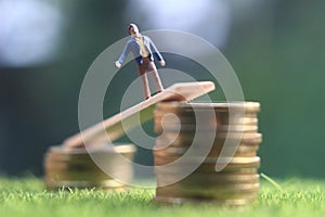 Miniature figure young bussinesman keep trying to get higher income walking at stack of coin at fresh green grass in the morning