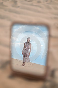 Photo in the mirror of attractive blond woman in long dress walking in desert.