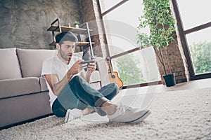 Photo of mixed race guy sitting floor leaning sofa holding telephone reading unexpected news not believe eyes wear