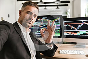 Photo of modern man taking selfie while working in office on computer with graphics and charts at screen