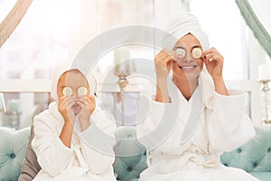 Photo of mother and daughter in white bathrobes. They applying pieces of cucumber to their eyes.