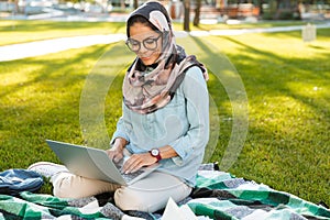 Photo of muslim female student 20s wearing headscarf sitting on blanket in green park