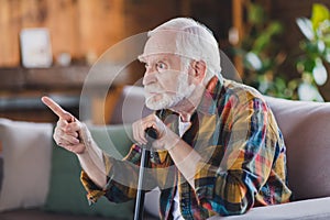 Photo of nervous angry retired man dressed plaid shirt holding nursery cane pointing finger sitting couch indoors