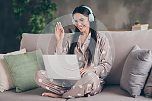 Photo of nice optimistic girl sit talk laptop wave hand wear spectacles headphones pijama at home photo