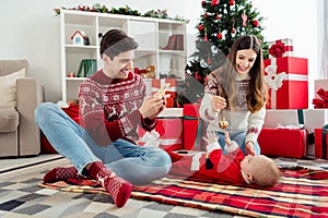 Photo of nice parents play little infant father hold telephone take selfie wear sweater in decorated x-mas home indoors