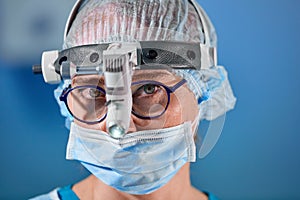 Photo of the operating surgeon in the surgery room. Surgeon in mask and glasses with mounted headlight. Close portrait