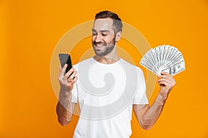 Photo of optimistic man 30s in casual wear holding cell phone and fan of money, isolated over yellow background