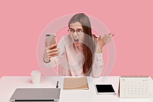 Photo of outraged annoyed woman holds modern cell phone, makes video call, argues with colleague, poses at workplace