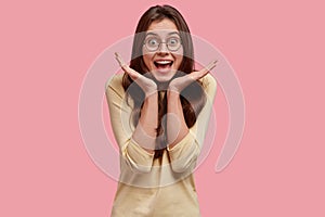 Photo of overjoyed glad woman keeps hands spread near face, happy and surprised to recieve good news, expresses