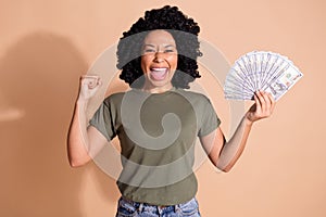 Photo of overjoyed woman with afro hairstyle wear khaki t-shirt hold money clenching fist win gambling isolated on beige