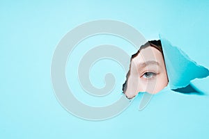 Photo of a painted eye of a girl looking at the camera through a hole on a blue background