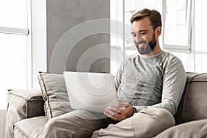 Photo of pleased adult man 30s in casual clothing typing on laptop, while working in cozy home