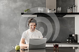 Photo of pleased adult man 30s in casual clothing typing on laptop, while working in headphones indoor