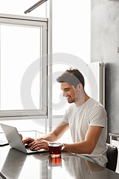 Photo of pleased adult man 30s in casual clothing typing on laptop, while working in modern apartment