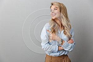 Photo of pleased blond businesswoman with long curly hair smiling and pointing finger aside at copyspace