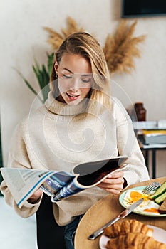 Photo of pleased blonde woman reading magazine while having breakfast