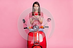 Photo portrait of amazed woman sitting on red moped using smartphone whistling isolated on pastel pink color background