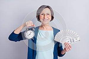 Photo portrait of cheerful business woman showing alarm clock keeping dollar banknotes isolated on grey color background