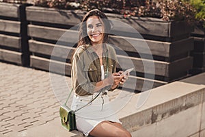 Photo portrait of cheerful girl holding phone in two hands sitting outside cafe waiting for friends to come