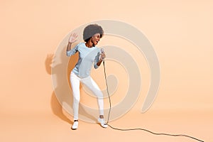 Photo portrait of dark skin woman singing in karaoke holding mic in one hand isolated on pastel beige colored background