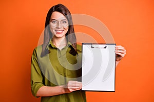 Photo portrait of female student keeping clipboard with white paper showing blank space isolated on vibrant orange color