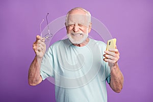 Photo portrait of happy elderly man taking off glasses celebrating victory holding phone in one hand isolated on vivid