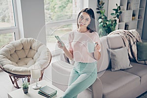 Photo portrait of happy young girl holding phone cup sitting on sofa arm relaxing indoors