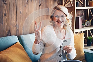 Photo portrait of lovely retired woman sit sofa device hold v-sign dressed casual outfit cozy home interior living room