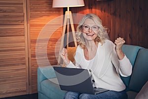 Photo portrait of lovely retired woman sit sofa laptop raise fists dressed casual outfit cozy home interior living room