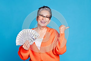 Photo portrait of lovely senior lady hold money fan point empty space dressed stylish orange garment isolated on blue