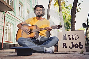Photo portrait of man performing on guitar sitting on street with carton looking for job black hat for money