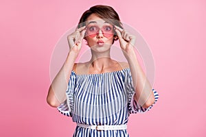 Photo portrait of pouting girl touching rose-tinted heart-shaped glasses  on pastel pink colored background