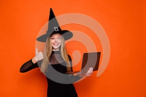 Photo portrait of pretty blonde teenager girl netbook showing thumb up dressed black halloween outfit isolated on orange