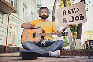 Photo portrait of sad grumpy man holding guitar sitting on street with carton looking for job headwear for money