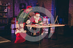 Photo portrait of santa claus reading green letter and elf putting big white gift box into present sack