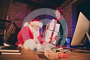 Photo portrait of santa claus writing with quill and ink in scroll and elf carrying three big presents