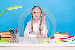 Photo portrait schoolgirl raising hand to answer on lesson sitting at desk isolated pastel blue color background