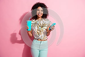 Photo portrait of smiling girl holding phone soft drink cup in hands isolated on pastel pink colored background