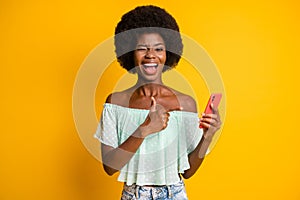 Photo portrait of winking girl holding phone in one hand pointing finger isolated on vivid yellow colored background