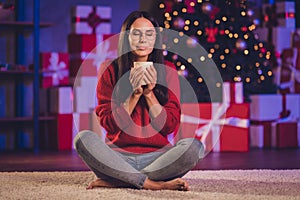 Photo portrait of woman smelling aroma of beverage holding mug in two hands sitting on floor in lotus pose indoors with