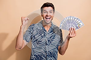 Photo portrait of young brunet hair guy raised fist up hold money to fly vacation lottery winner isolated on beige color
