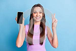 Photo portrait of young girl demonstrating mobile phone touchscreen copyspace showing v-sign isolated on vivid blue