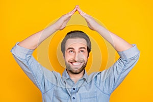 Photo portrait of young guy smiling keeping hands over head safety insurance looking up isolated on bright yellow color